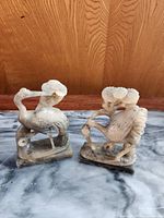 Two intricately carved stone crane sculptures side by side on marble surface, wooden background.