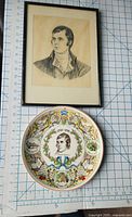 Framed portrait pencil drawing alongside ceramic commemorative Robert Burns plate on grid background for size reference.