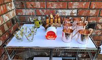 Photo showing entire barware lot including shot glasses, mini beer mug, cork stoppers, ashtray, coasters with caddy, and Smirnoff copper mugs arranged on a white table against brick wall.