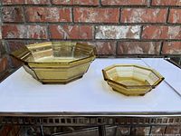 Two octagonal amber glass bowls placed side by side on a glass table with a brick wall background. Larger bowl on left, smaller on right, showing shape and texture.