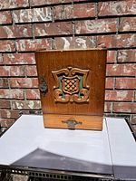 Front view of the vintage wooden storage cupboard showing the carved ornate door with shield decoration and metal latch handle.