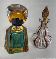 Photo of two art glass perfume bottles, one geometric in amber, green and blue with round stopper, one striped in white, pink and gold with leaf-shaped stopper