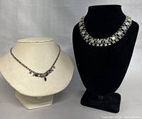 Two vintage signed costume jewelry necklaces displayed on bust forms: a silver-tone pyramid-shaped necklace on black bust and a rhinestone floral design necklace on light bust.