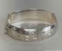 Silver engraved bangle bracelet shown from front angle with safety chain visible.
