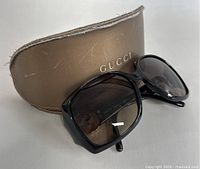 Black oversized Gucci sunglasses with gradient grey lenses and gold Gucci 'GG' logo on temples, positioned in front of the brown leather Gucci case with gold logo on the front. Case shows minor wear and scratches.