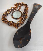 Both the 10-inch tortoiseshell serving spoon and the MCM acrylic magnifier pendant necklace lying flat to show both items in context.