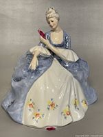 Front view of Royal Doulton "Wistful" figurine showing seated woman in blue and white dress with floral decorations and pink fan in hand.