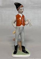 Front view of the Hollohaza porcelain figurine depicting a woodcutter boy, showing the full figure, orange vest, saw in hand, and small wood stump at feet.