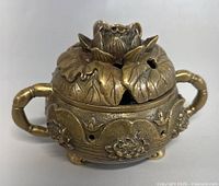 Front angled view showing detailed lotus flower lid with perforations and one handle in view, resting on three small feet. The copper metal has a patinated vintage look.