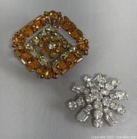 Both brooches shown with full view of the orange square rhinestone brooch and the clear starburst brooch side by side