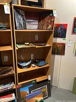 Bookshelf in faux light wood tone with 5 shelves. Includes various paintings and ceramic pieces placed on and around it for display only, not included.
