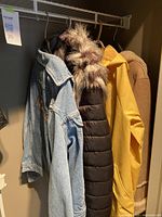 Four ladies coats hanging in closet: light blue denim jacket, black puffer coat with faux fur, yellow raincoat, and tan suede shearling coat