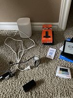 Lot items arranged on carpet: white Sonos speaker, Amazon Fire TV Stick 4K box, Kindle with open cover, charging cords, and thermostat device