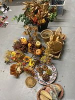 Assortment of fall decorative items including candles, floral picks, baskets, and faux foliage planter in background