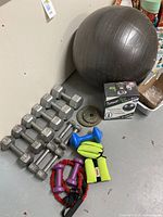 Photo showing a variety of dumbbells from 3 to 20 pounds along with a boxed Forever Flex stability ball, resistance bands, wrist weights, and other hand weights arranged on a gray floor.