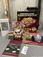 Photo showing boxed Solid Wood & Brass 6 Game Ensemble alongside Jiggy Animal Kingdom puzzle, Golden Girls puzzle, Scrabble, various boxed games, Cribbage book, and a bag of game pieces.