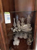 Wide view of glassware assortment on glass shelf in wooden cabinet, showing multiple pieces including vases, decanter, candy dish, candle holders and glasses.