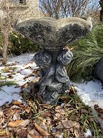 Full view of birdbath showing scalloped basin and cherub base from the front outdoors on ground with leaves and snow.