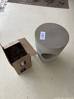 Photo shows small round grey outdoor table and adjacent cardboard box with plastic wine glasses inside on a concrete floor.