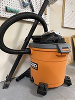 Full side view of orange Ridgid 60 litre wet/dry vacuum with hose attached, nozzle connected, showing caster wheels and orange body with black top.