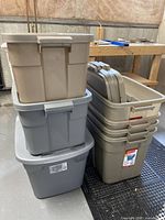 Seven storage totes stacked in a basement setting on black interlocking tiles. Four larger beige Rubbermaid totes on the right, with four lids visible. Three stacked gray and beige totes on the left.