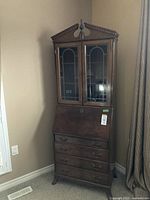 Full view of the wood secretary cabinet in a corner, showing the glass-front upper section, drop-down desk, and four drawers below.