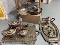 Overall view of multiple silver plated serveware items including coffee and tea pots, cream and sugar bowls, large ornate tray, and additional trays with gravy boat.