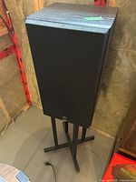 Front view of one speaker mounted on a black metal stand showing cabinet and grille.