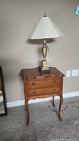 Wooden scalloped top side table with two drawers and metal base table lamp with linen shade on carpeted floor.