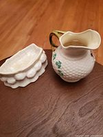 Wide view of both Belleek porcelain pieces displayed on a wooden surface, showing the rowboat dish and the basket-shaped shamrock decorated piece