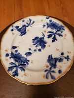 Top view of one antique porcelain plate showing blue floral decoration and gold trim, with visible age spots and stains on surface.