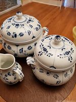Three porcelain pieces including two covered pots and a small pitcher with blue floral patterns on a wooden floor.