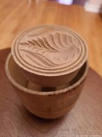 Top and side view of antique wooden butter press showing carved wheat and leaf designs on lid.