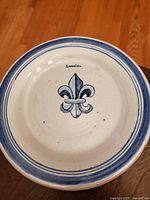 Front view of a round ceramic plate featuring a fleur-de-lis symbol painted in blue in the center and bordered by blue rings.