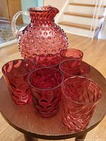 Photo of the antique cranberry hobnail pitcher with six matching glasses arranged on a wooden table, showing the hobnail texture and cranberry color clearly.