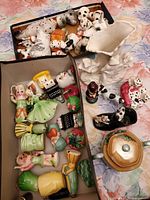 Top view photo showing two boxes of assorted vintage Japanese trinkets including various animals, characters, and decorative ceramics on floral tablecloth.