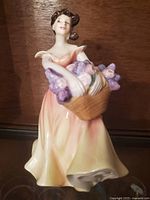 Front view of the Royal Doulton Lesley figurine showing a woman in peach and cream dress holding a basket of purple flowers.
