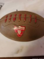 Close-up photo of brown football-shaped musical wind-up toy showing Harvard crest with red shield and text, with textured surface and red molded laces on top.