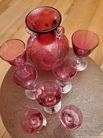 Photo showing the vintage cranberry decanter surrounded by six glasses on a wooden round surface, highlighting the rich cranberry red color and clear stems of the glasses.