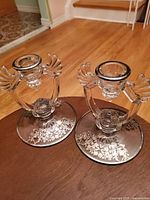 Pair of vintage glass candle holders shown on wooden surface with detailed sterling silver overlay design visible on bases and wing-shaped glass accents on each side.