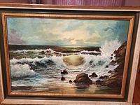Framed oil painting showing crashing ocean waves and rocky shore under cloudy sky with sunlight.