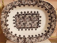 Full view of oval ceramic serving platter showing intricate black lace-like floral and starburst transferware pattern on white ceramic base.