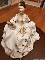 Front view of the white porcelain figurine of a woman seated in a flowing dress with gold leaf details and pink flowers, showing overall condition and design.