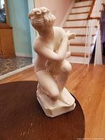 Full view of the sculpture showing the crouching Venus figure from a front angle on a wooden surface with interior background.