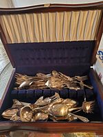 Wooden box with fabric lining containing assorted vintage forks, spoons, and cutting utensils piled inside the slot sections