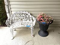 White cast metal bench next to black plastic planter with pink artificial flowers on concrete floor against house siding.