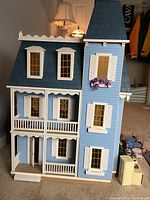 Front view of the handcrafted wooden dollhouse showing blue paint, white shutters, multiple windows, and a shingled roof. Dollhouse stands with some accessories nearby.