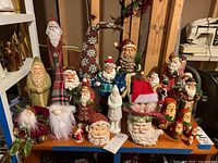 Overview of multiple Santa Claus figurines of different sizes, materials, and styles displayed on a wooden shelf.
