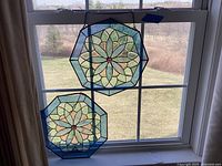 Two octagonal stained glass sun catchers hanging in a window with a scenic background, showing green and yellow glass petals with red central pieces and blue border frames.