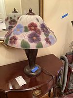 Full view of the table lamp showing painted glass flower shade and metal base on wooden furniture.